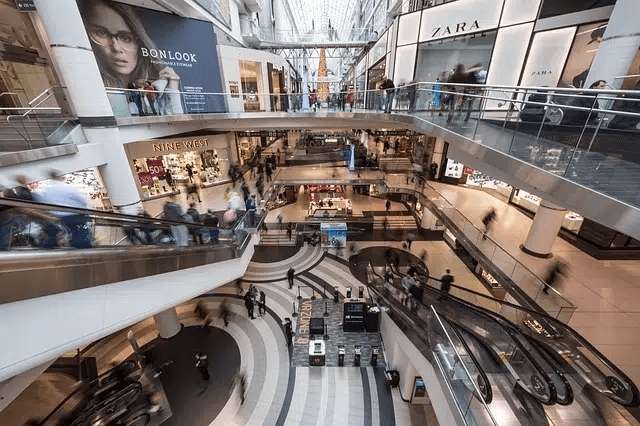 Gente moviéndose vertiginosamente en una plaza de una tienda a otra. La fotografía desenfoca lo que se mueve para dar una sensación de velocidad.